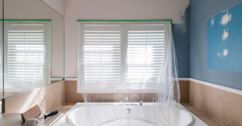 Bathroom in the process of being painted, plastic and tape over window above bathtub.