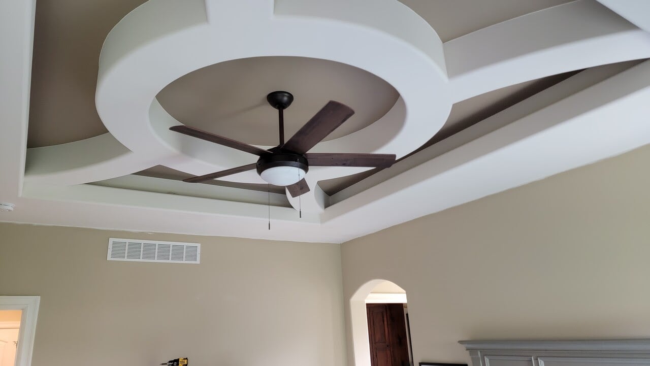 Bedroom ceiling painted light brown same color as the walls. There is white bordered molding around dark brown colored ceiling fan.