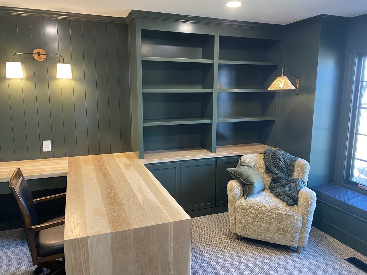 Interior home office with a built in desk and bookshelf painted dark emerald green with light wood desk.