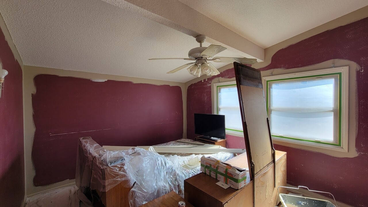 Interior bedroom with furniture covered in plastic and maroon painted wall being painted beige with paint brushed along top of wall near ceiling and in corners.