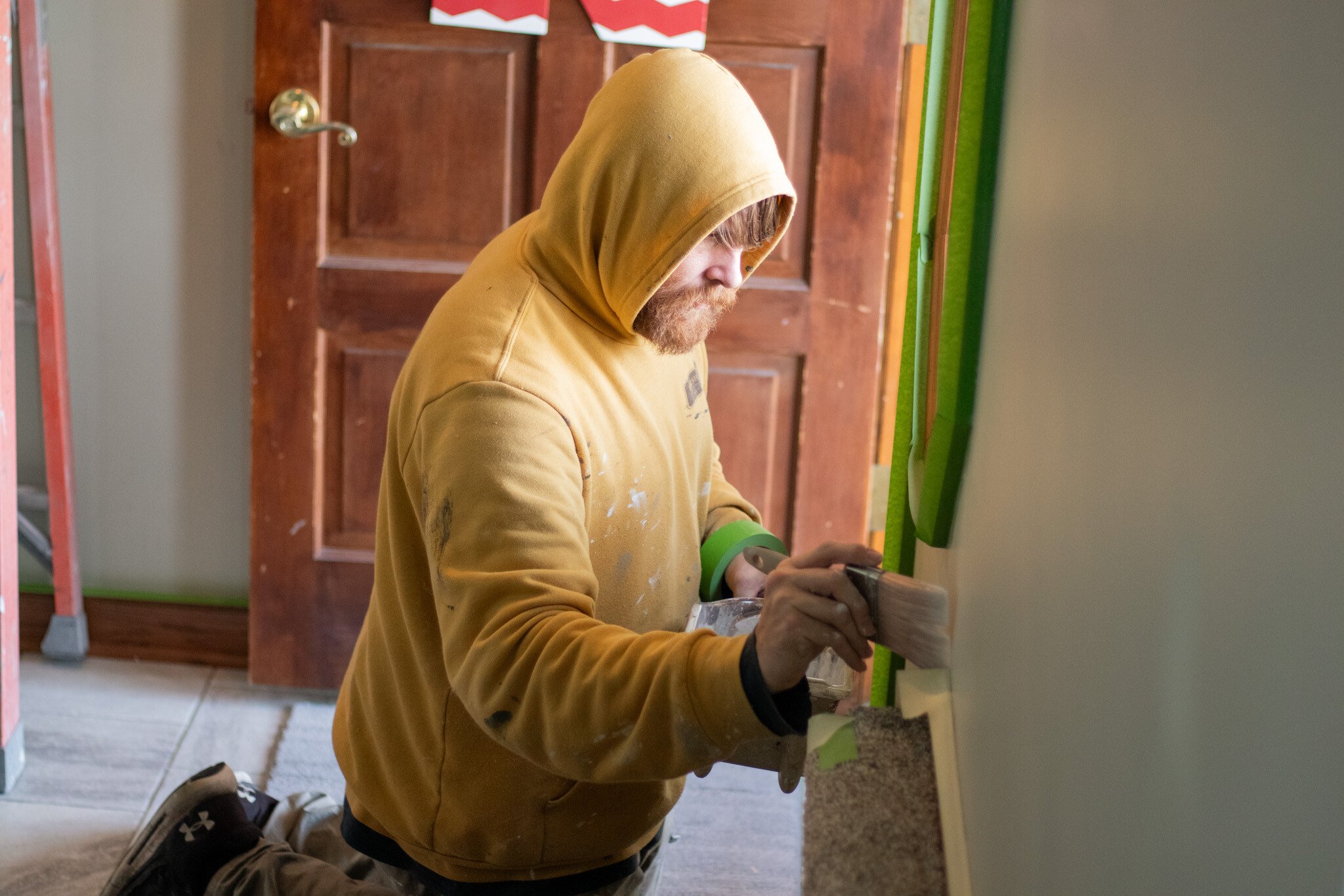 Painter brushing paint on a wall near the front door of a home with tape along edges.