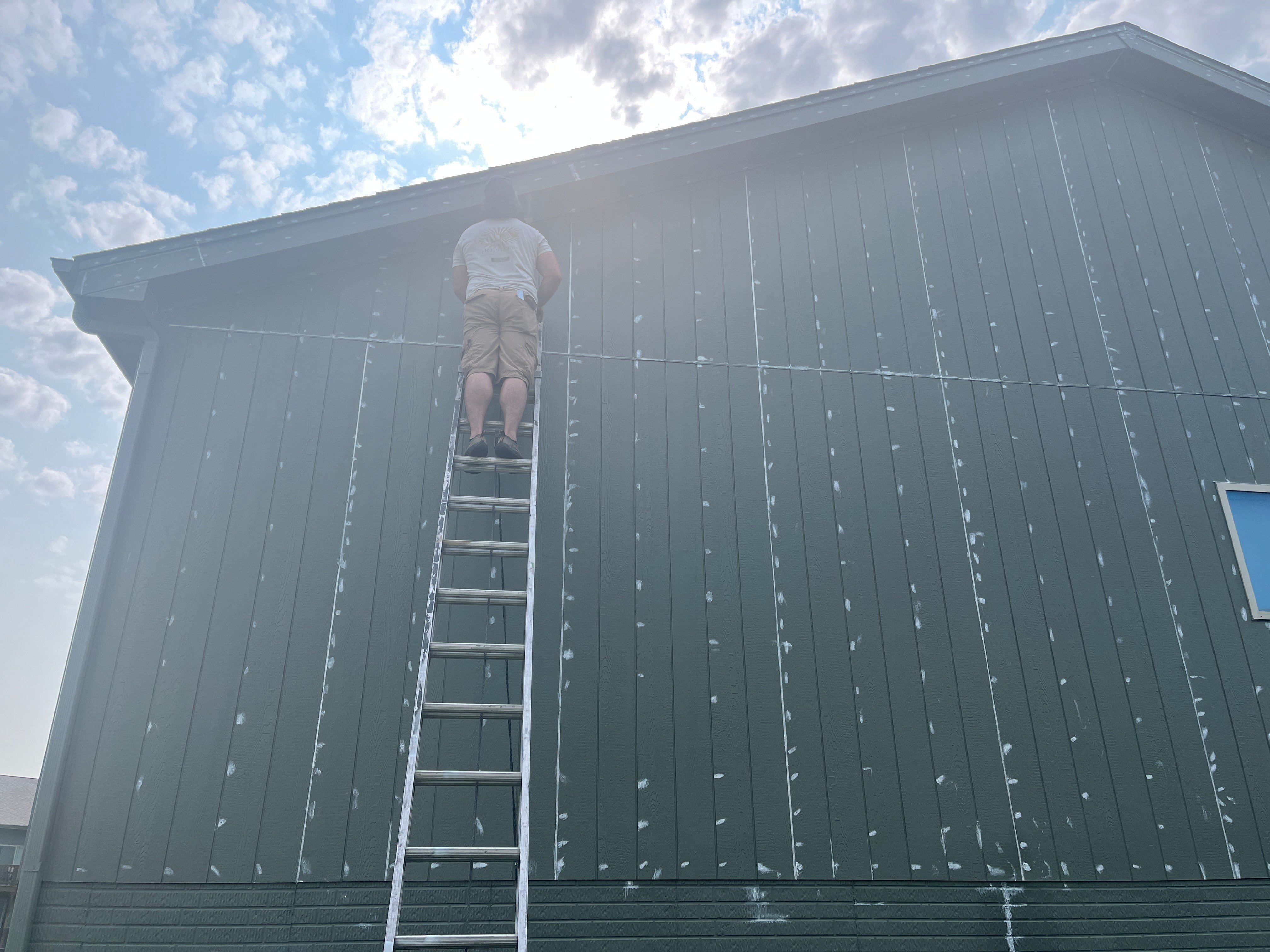 Painter on a ladder against the side of dark green home in Omaha, caulking gaps in the siding.