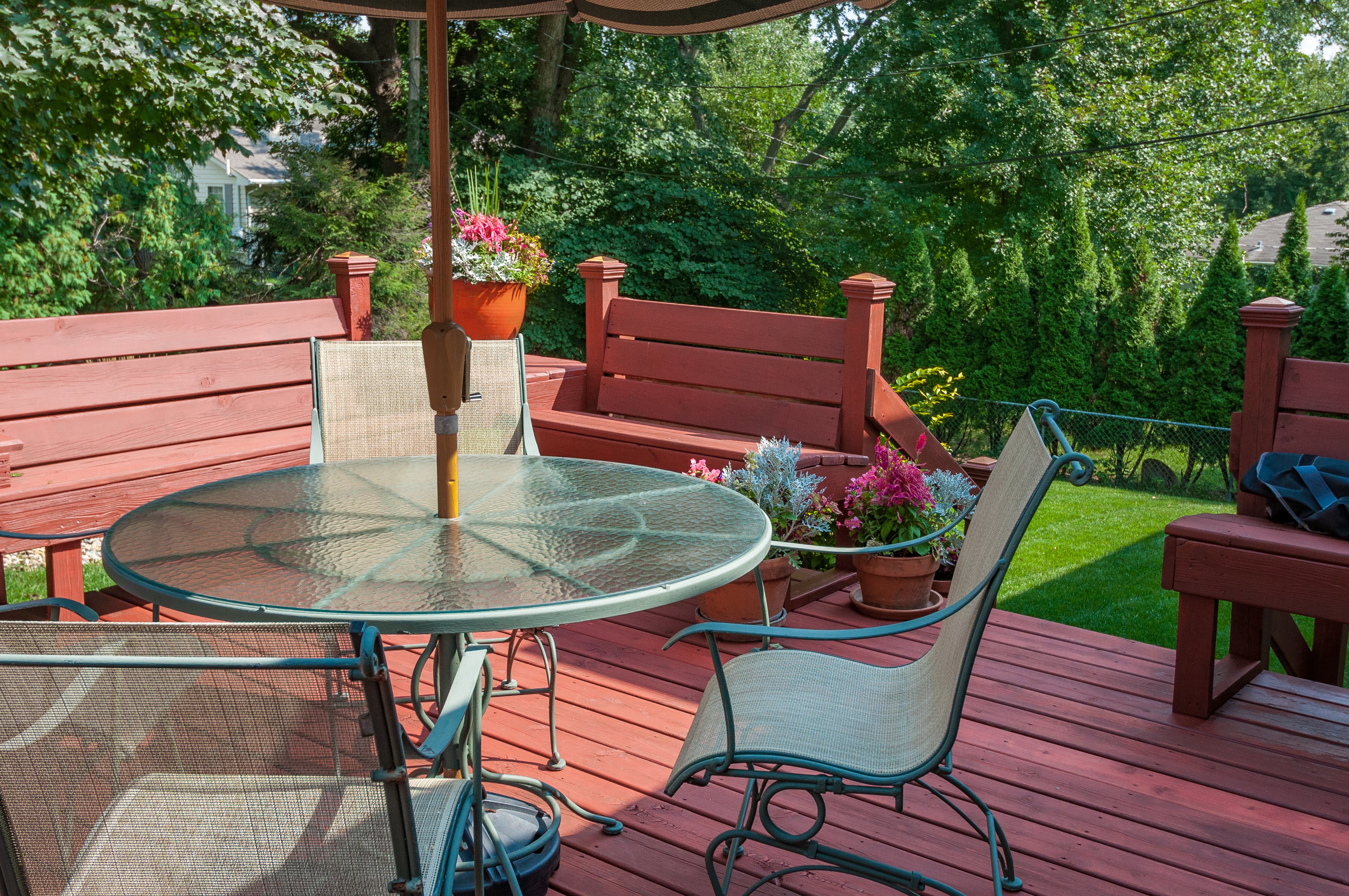 Redish-brown stained deck with a glass and black metal table and chair set on top. Flowers in pots on the deck.