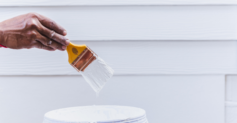 Man dipping paint brush into white paint, about to paint siding on a home white.